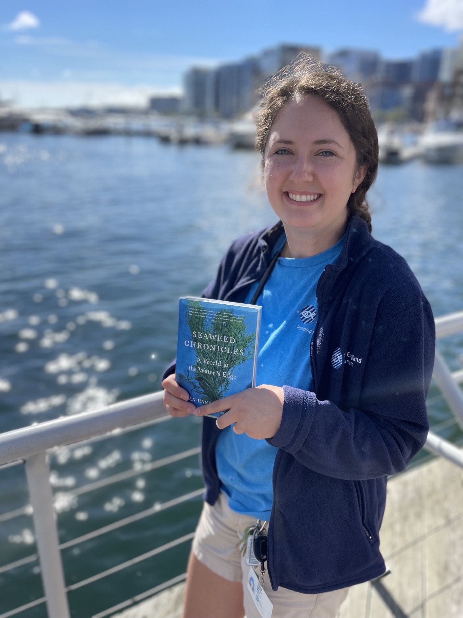 Jordan holding a copy of Seaweed Chronicles standing in front of the harbor