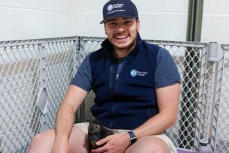 Tommy is sitting on the ground and a penguin chick is in his lap. He's smiling at the camera.