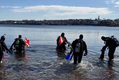 Five scuba divers entering the sea carrying diving equipment and a diver down flag.