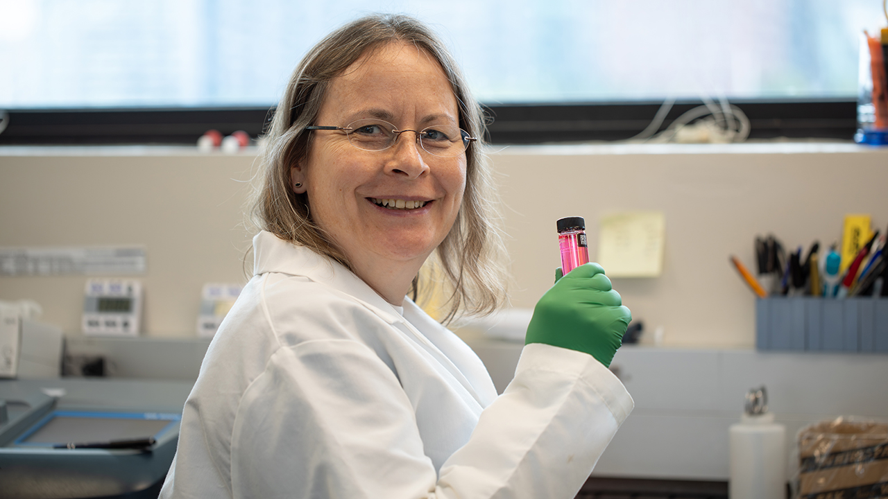 A person in a lab coat smiling at the camera and holding a test tube filled with red liquid