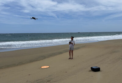 A woman operates a drone on a sandy beach, with a round landing pad nearby and a black carrying case on the sand.