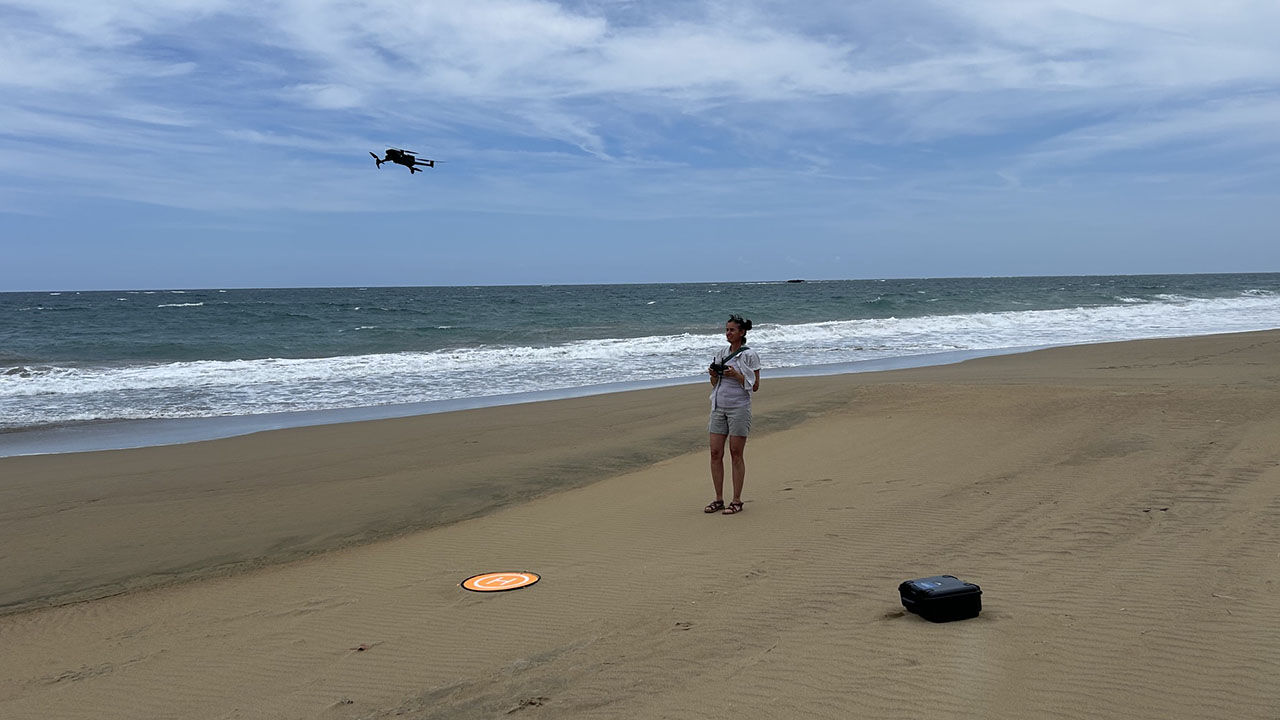 A woman operates a drone on a sandy beach, with a round landing pad nearby and a black carrying case on the sand.