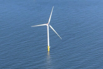 Offshore wind turbine with three white blades and a yellow base standing in blue water.