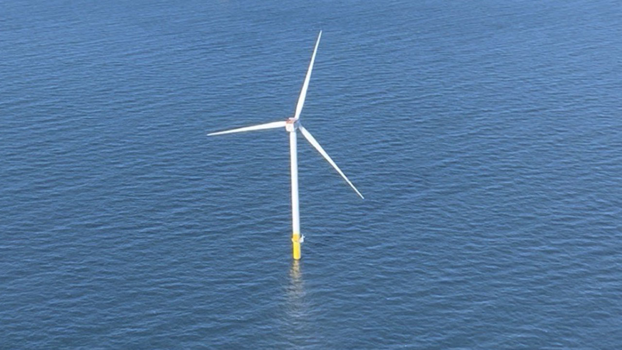Offshore wind turbine with three white blades and a yellow base standing in blue water.