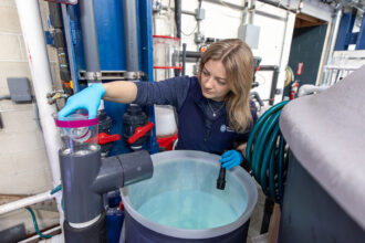A woman in blue gloves inspects a jar above a round container of clear liquid in a laboratory filled with pipes and valves