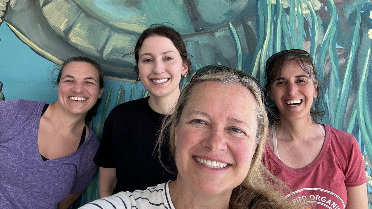 Four women smiling together in front of a colorful mural featuring abstract plants in teal and green hues.