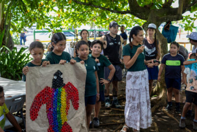 A community gathering with children holding a poster displaying a sea turtle made out of colorful bottle caps.
